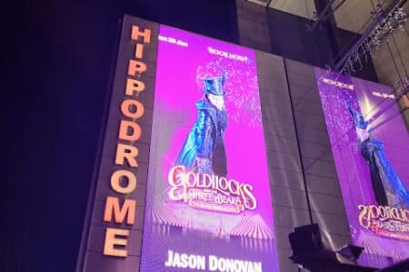 A large illuminated sign outside the Hippodrome theatre at night displays a bright purple poster for “Goldilocks and the Three Bears,” featuring a performer in a top hat and long coat. The name “Jason Donovan” appears at the bottom of the poster.