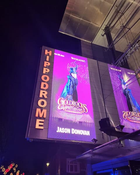 A large illuminated sign outside the Hippodrome theatre at night displays a bright purple poster for “Goldilocks and the Three Bears,” featuring a performer in a top hat and long coat. The name “Jason Donovan” appears at the bottom of the poster.