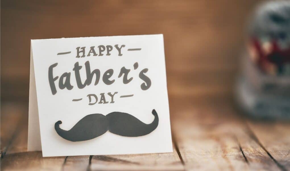 A white greetings card on a wooden surface reads "Happy Father's Day" in bold, playful letters, with a black moustache graphic below the text. The background is softly blurred, highlighting the card as the main focus.