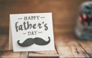 A white greetings card on a wooden surface reads "Happy Father's Day" in bold, playful letters, with a black moustache graphic below the text. The background is softly blurred, highlighting the card as the main focus.