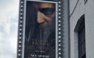 A vertical marquee sign on a white building displays a poster for "Derren Brown: Showman Live Tour" at The Alexandra. The poster shows Derren Brown’s intense face with text about the event dates: Tue 6 – Sat 10 Sept, and a website link for tickets.
