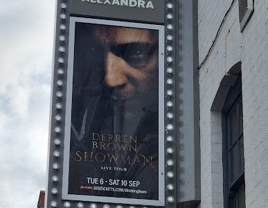 A vertical marquee sign on a white building displays a poster for "Derren Brown: Showman Live Tour" at The Alexandra. The poster shows Derren Brown’s intense face with text about the event dates: Tue 6 – Sat 10 Sept, and a website link for tickets.