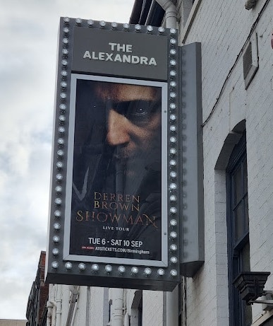 A vertical marquee sign on a white building displays a poster for "Derren Brown: Showman Live Tour" at The Alexandra. The poster shows Derren Brown’s intense face with text about the event dates: Tue 6 – Sat 10 Sept, and a website link for tickets.