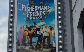 A vertical marquee sign for The Alexandra theatre displays a poster for “Fisherman’s Friends: The Musical.” The poster shows cheerful people with fishing gear and musical instruments on a dock, with text praising the musical and dates at the bottom.