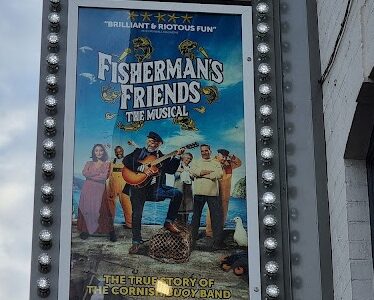 A vertical marquee sign for The Alexandra theatre displays a poster for “Fisherman’s Friends: The Musical.” The poster shows cheerful people with fishing gear and musical instruments on a dock, with text praising the musical and dates at the bottom.