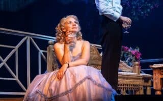 A woman in a pink ballgown sits on a bench holding a glass, gazing upwards. Behind her, a man in a dinner jacket stands with a wine glass, looking into the distance. The stage is set with elegant furniture and blue-purple theatrical lighting.