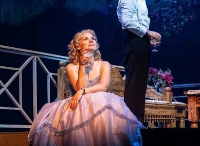 A woman in a pink ballgown sits on a bench holding a glass, gazing upwards. Behind her, a man in a dinner jacket stands with a wine glass, looking into the distance. The stage is set with elegant furniture and blue-purple theatrical lighting.