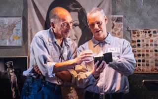 Two men in prison uniforms stand close together in a dimly lit cell, smiling as they look at a small black box with light shining from it. Behind them are posters and a map on the wall, including a large image of a woman’s face and black-and-white photographs.
