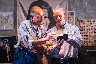 Two men in prison uniforms stand close together in a dimly lit cell, smiling as they look at a small black box with light shining from it. Behind them are posters and a map on the wall, including a large image of a woman’s face and black-and-white photographs.