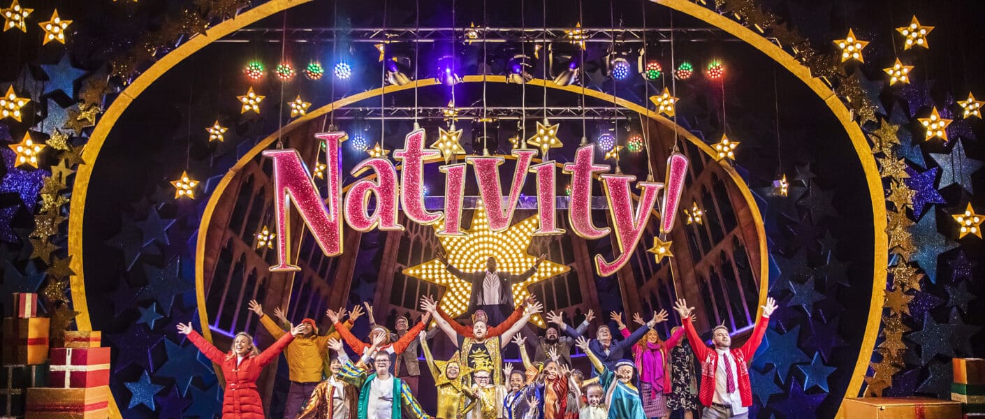 A large group of children and adults in colourful costumes stand on stage, arms raised in excitement, beneath a glittering sign that reads “Nativity!” with a large gold star. The festive stage is decorated with bright lights, gift boxes, and glowing star-shaped ornaments.