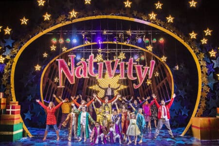 A large group of children and adults in colourful costumes stand on stage, arms raised in excitement, beneath a glittering sign that reads “Nativity!” with a large gold star. The festive stage is decorated with bright lights, gift boxes, and glowing star-shaped ornaments.