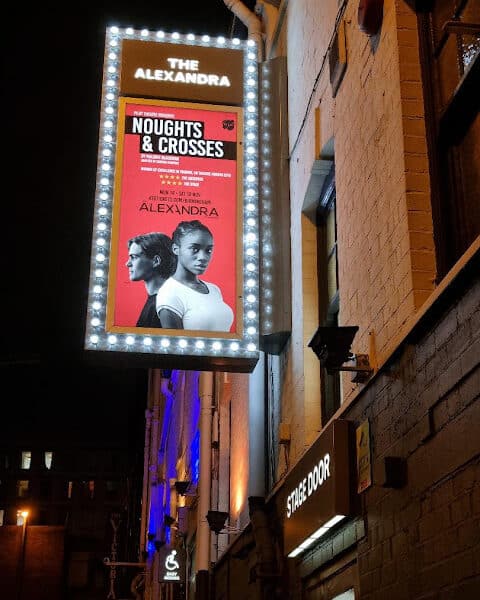 A lit theatre marquee for The Alexandra displays a poster for "Noughts & Crosses." The red poster shows a serious-looking man and woman standing back-to-back, along with show details in white text. The sign is bordered by bright lights.