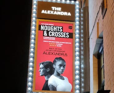 A lit theatre marquee for The Alexandra displays a poster for "Noughts & Crosses." The red poster shows a serious-looking man and woman standing back-to-back, along with show details in white text. The sign is bordered by bright lights.