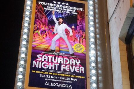 A brightly lit marquee for The Alexandra theatre displays a poster for "Saturday Night Fever." The poster features a man in a white suit dancing on a colourful disco floor with a lively crowd and spotlights in the background. Dates and show info are listed below.