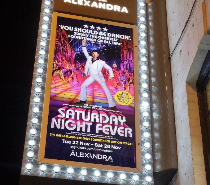 A brightly lit marquee for The Alexandra theatre displays a poster for "Saturday Night Fever." The poster features a man in a white suit dancing on a colourful disco floor with a lively crowd and spotlights in the background. Dates and show info are listed below.
