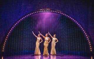 Three women in sparkling gold gowns stand centre stage under a glowing arch. Each strikes a dramatic pose with one arm raised. Purple stage lights and a shimmering curtain create a glamorous, showbiz atmosphere.