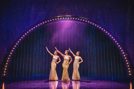 Three women in sparkling gold gowns stand centre stage under a glowing arch. Each strikes a dramatic pose with one arm raised. Purple stage lights and a shimmering curtain create a glamorous, showbiz atmosphere.