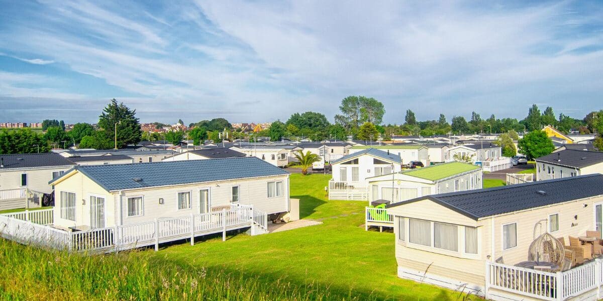 Rows of modern, white mobile homes with dark roofs are set on bright green lawns. Each home has a small porch or decking. The sky above is blue with wispy clouds, and trees and buildings are visible in the distant background.
