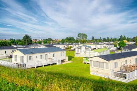 Rows of modern, white mobile homes with dark roofs are set on bright green lawns. Each home has a small porch or decking. The sky above is blue with wispy clouds, and trees and buildings are visible in the distant background.