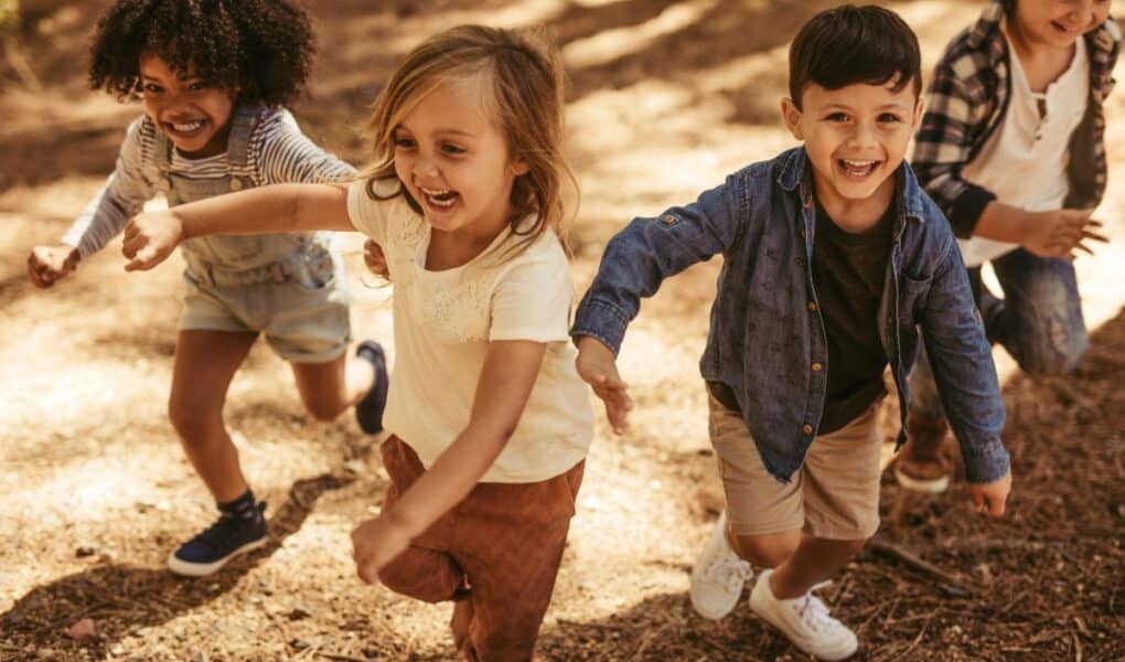 Four young children laugh and run outdoors on a sunny day. They wear casual summer clothes and appear joyful as they play together on a dirt and pine needle-covered ground, with trees and dappled sunlight in the background.