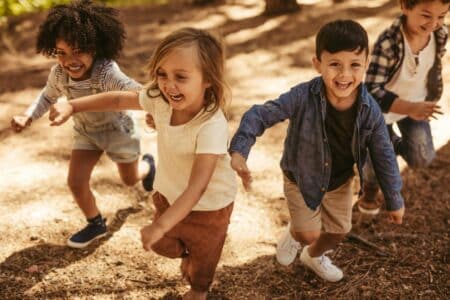 Four young children laugh and run outdoors on a sunny day. They wear casual summer clothes and appear joyful as they play together on a dirt and pine needle-covered ground, with trees and dappled sunlight in the background.