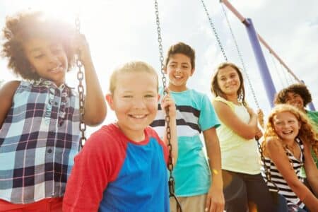 A group of six smiling children of diverse backgrounds stand together outdoors on swings. They are dressed in bright, casual summer clothes and bathed in sunlight, with a playground swing set and blue sky in the background.