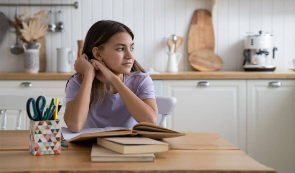 A girl with brown hair sits at a wooden table, resting her chin on her hands and gazing to the side. In front of her are open and closed books, with a colourful cup of pens nearby. The background shows a bright kitchen with white cupboards and wooden decor.