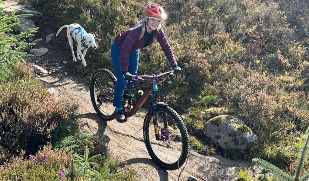 A woman wearing a red helmet and sunglasses rides a mountain bike along a narrow dirt track through grassy terrain. A white Dalmatian dog with black spots runs behind her, following on the path. Sunlight brightens the scene and wild plants line the track.