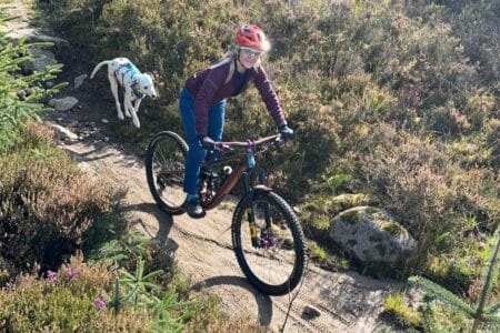 A woman wearing a red helmet and sunglasses rides a mountain bike along a narrow dirt track through grassy terrain. A white Dalmatian dog with black spots runs behind her, following on the path. Sunlight brightens the scene and wild plants line the track.