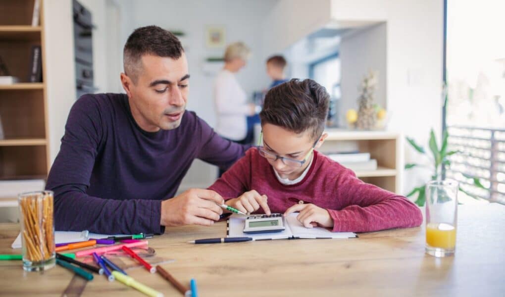 A man helps a boy with homework at a wooden table covered in coloured markers, pencils, and paper. The boy writes in a notebook while the man points. In the background, two adults talk in a bright kitchen. A glass of orange juice sits on the table.