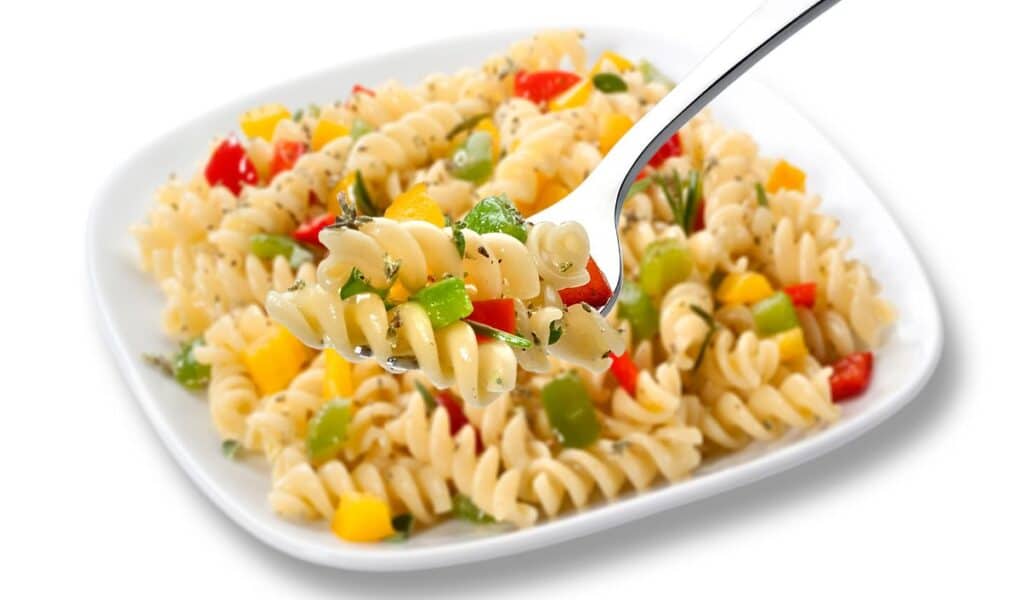 A square white plate holds rotini pasta mixed with diced yellow, red, and green peppers, and herbs. A fork in the foreground lifts a serving, showing the colourful vegetables and spiral pasta in detail.