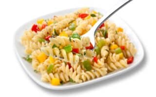 A square white plate holds rotini pasta mixed with diced yellow, red, and green peppers, and herbs. A fork in the foreground lifts a serving, showing the colourful vegetables and spiral pasta in detail.