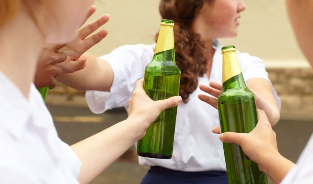 Three young people in white shirts hold green glass beer bottles outdoors. One person turns away, whilst others gesture with their hands, as if refusing or pushing the bottles away. The background is blurred but shows a stone wall and pavement.