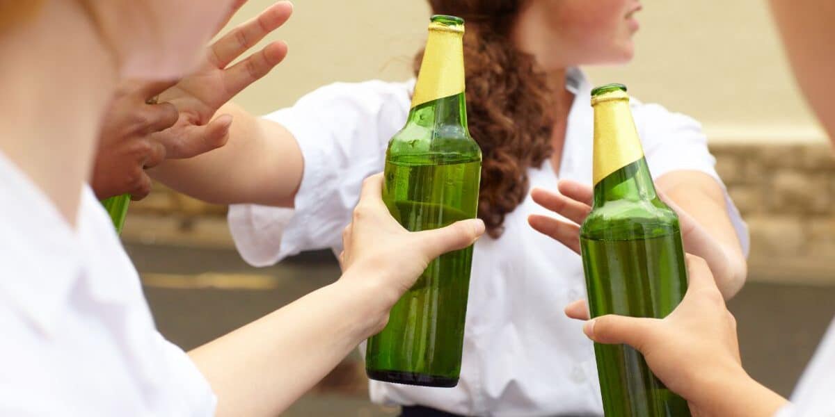 Three young people in white shirts hold green glass beer bottles outdoors. One person turns away, whilst others gesture with their hands, as if refusing or pushing the bottles away. The background is blurred but shows a stone wall and pavement.