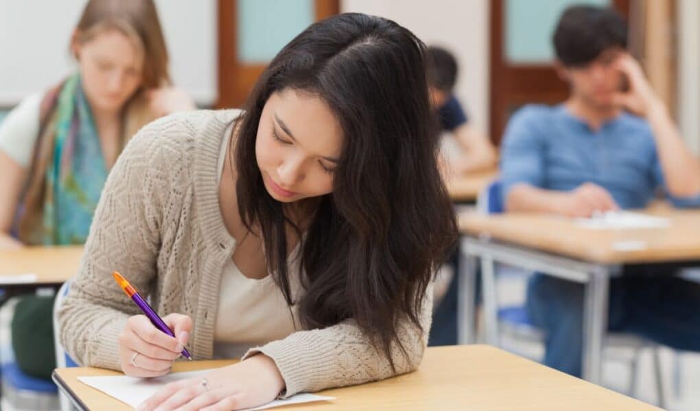 A young woman with long dark hair writes on paper at a desk in a classroom, focusing intently. She wears a tan cardigan. Other students sit at desks in the background, also working quietly. The classroom has light-coloured walls and wooden doors.