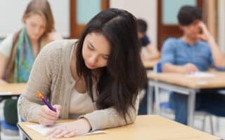 A young woman with long dark hair writes on paper at a desk in a classroom, focusing intently. She wears a tan cardigan. Other students sit at desks in the background, also working quietly. The classroom has light-coloured walls and wooden doors.