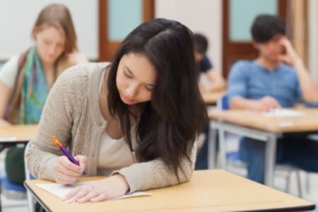 A young woman with long dark hair writes on paper at a desk in a classroom, focusing intently. She wears a tan cardigan. Other students sit at desks in the background, also working quietly. The classroom has light-coloured walls and wooden doors.