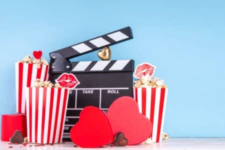 A film clapperboard sits among red-and-white striped popcorn tubs, red heart shapes, and chocolates against a light blue background. Popcorn is decorated with red lips and heart toppers, creating a festive, romantic film night setup.