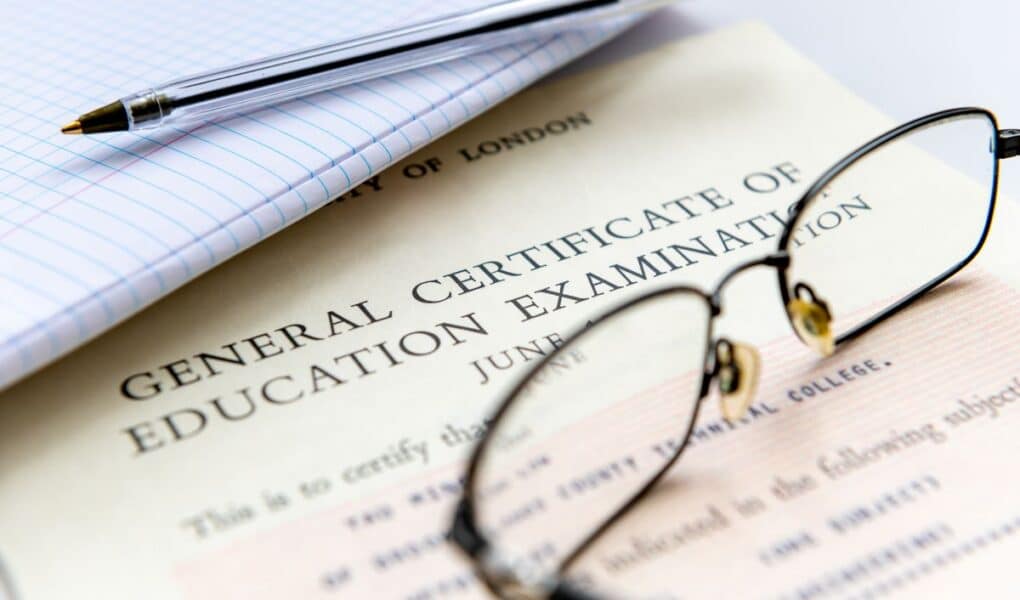 Close-up of a GCE (General Certificate of Education) certificate partly covered by spectacles, a pen, and a lined notebook, suggesting an academic or exam-related setting. The certificate heading and part of the date are visible.