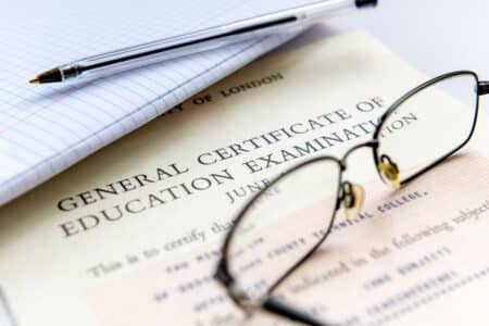 Close-up of a GCE (General Certificate of Education) certificate partly covered by spectacles, a pen, and a lined notebook, suggesting an academic or exam-related setting. The certificate heading and part of the date are visible.