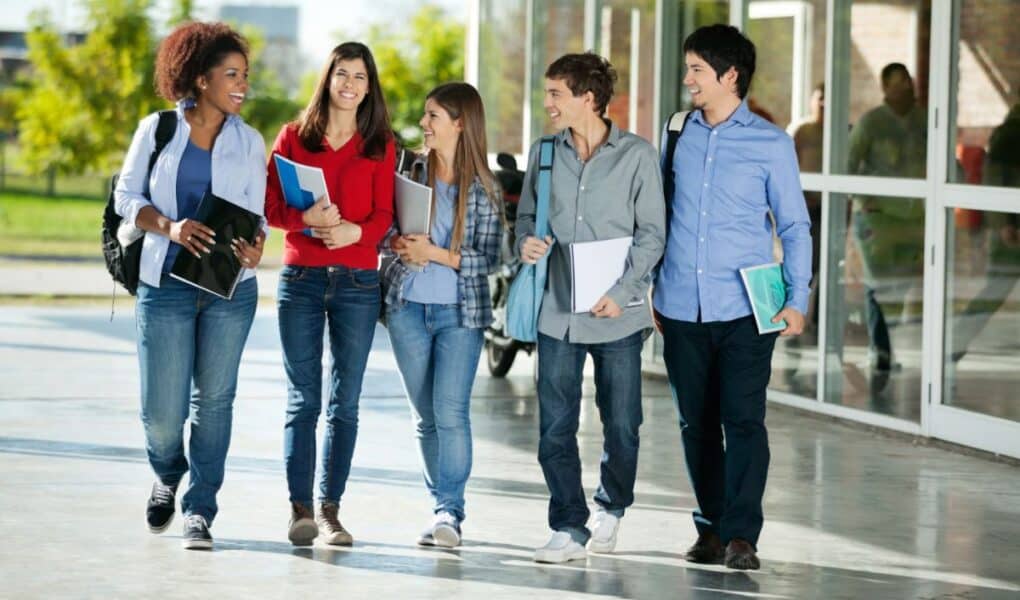 Five students walk together outside a modern building, smiling and chatting. They wear casual clothes and carry rucksacks and notebooks, appearing relaxed and friendly. Trees and reflections are visible in the background.