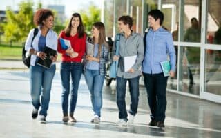Five students walk together outside a modern building, smiling and chatting. They wear casual clothes and carry rucksacks and notebooks, appearing relaxed and friendly. Trees and reflections are visible in the background.
