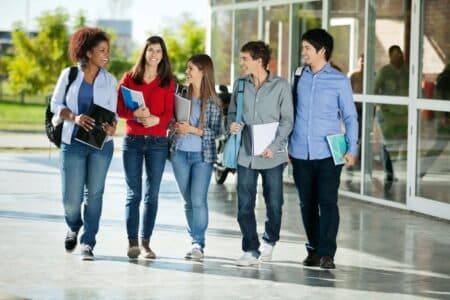 Five students walk together outside a modern building, smiling and chatting. They wear casual clothes and carry rucksacks and notebooks, appearing relaxed and friendly. Trees and reflections are visible in the background.