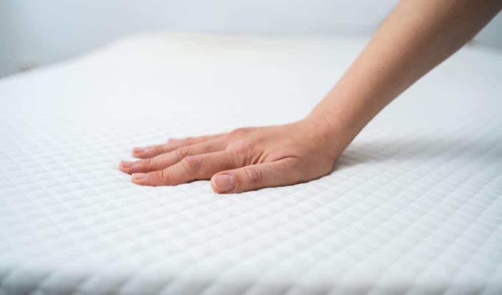 A hand presses down on a white mattress with a quilted diamond pattern, testing its firmness. The background is blurred, focusing attention on the hand and mattress surface.
