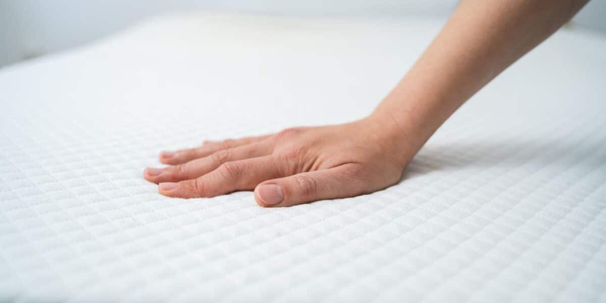 A hand presses down on a white mattress with a quilted diamond pattern, testing its firmness. The background is blurred, focusing attention on the hand and mattress surface.