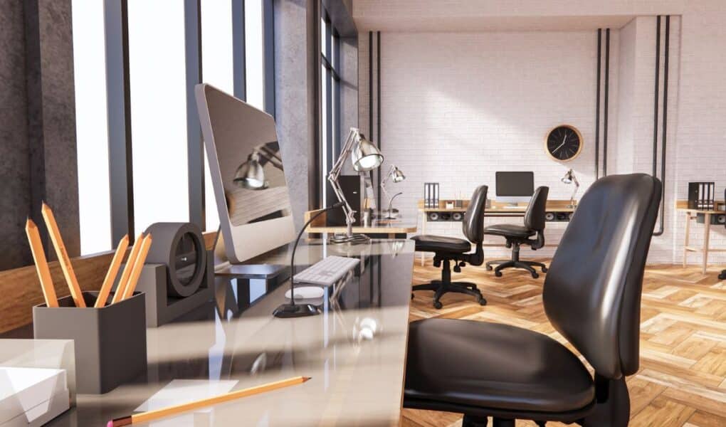 A modern open-plan office with several desks, computers, office chairs, desk lamps, and pencil pots. Large windows let in natural light, and there is a black wall clock on a white brick wall in the background. The floor is wood-patterned.