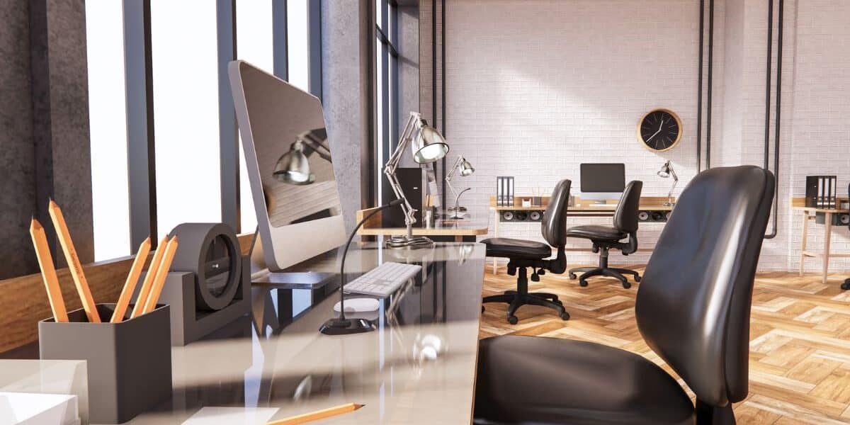 A modern open-plan office with several desks, computers, office chairs, desk lamps, and pencil pots. Large windows let in natural light, and there is a black wall clock on a white brick wall in the background. The floor is wood-patterned.