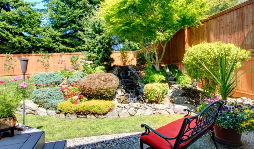 A landscaped back garden features a small waterfall surrounded by rocks, colourful bushes, and green trees. A wooden fence encloses the garden. In the foreground, a black bench with a red cushion sits on a gravel patio next to potted flowering plants.