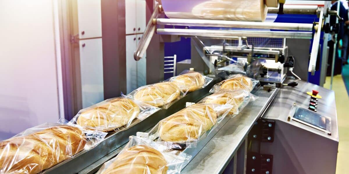 A packaging machine wraps several loaves of bread in transparent plastic film on a conveyor belt. The freshly packed loaves move in a row through the automated system inside an industrial food production facility.