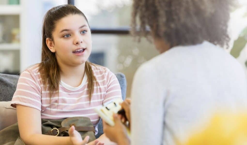 A teenage girl with brown hair in a ponytail, wearing a pink striped shirt, sits on a sofa and talks to a woman holding a notepad, seen from behind. The setting appears to be a casual, well-lit counselling or therapy session.